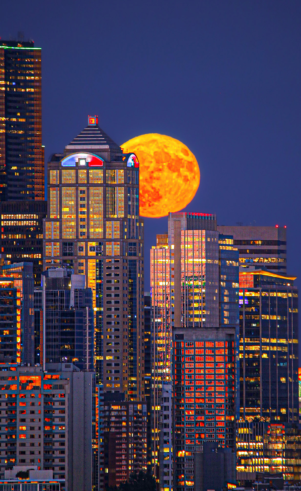 This picture reveals the city of Seattle, illuminated at twilight during a summer sunset with the orange Buck Moon rising among the skyline
